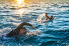 Dos nadadores practicando estilo libre en aguas abiertas al atardecer, entre olas y salpicaduras iluminadas por el sol.