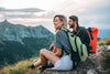 Dos excursionistas con mochilas descansan en una montaña mientras observan el paisaje al atardecer.