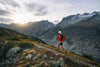 Corredor practicando trail running por un sendero rocoso de montaña, rodeado de vegetación y picos montañosos al atardecer