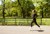 Una mujer con ropa deportiva negra corre por un sendero de un parque soleado y rodeado de árboles verdes.