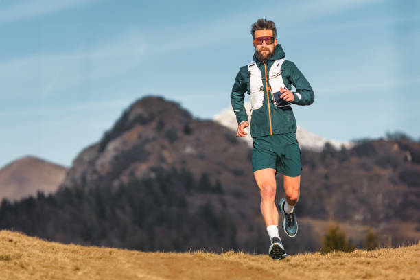 Hombre corriendo por un sendero al aire libre en una zona montañosa, con ropa deportiva y gafas de sol, rodeado de naturaleza durante un día despejado