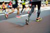 Corredores participando en una carrera de larga distancia por una avenida, con enfoque en sus piernas y tenis deportivos en movimiento.