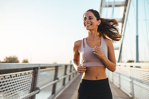 Mujer sonriente corriendo al aire libre sobre un puente, vestida con ropa deportiva, durante un entrenamiento en un día soleado.